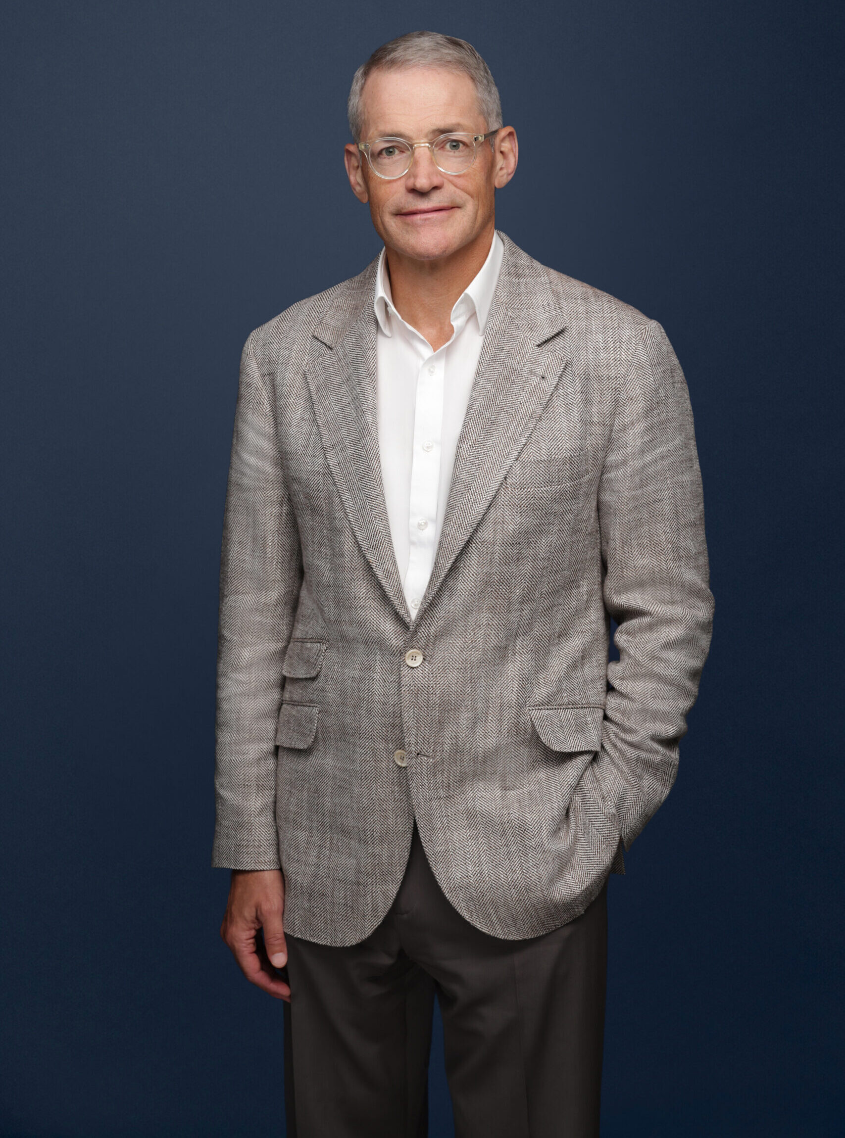 Medium long shot of Michael Fisch wearing a light grey blazer, white button-down, and taupe pants against a navy blue backdrop.